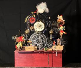 Machine avec rouages, horloge, lutins miniatures, fils, boîtes, le tout sur un socle rouge. Vue de face