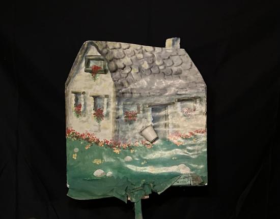 Maisonnette dans un pré, avec des fleurs et un bidon blanc devant la porte