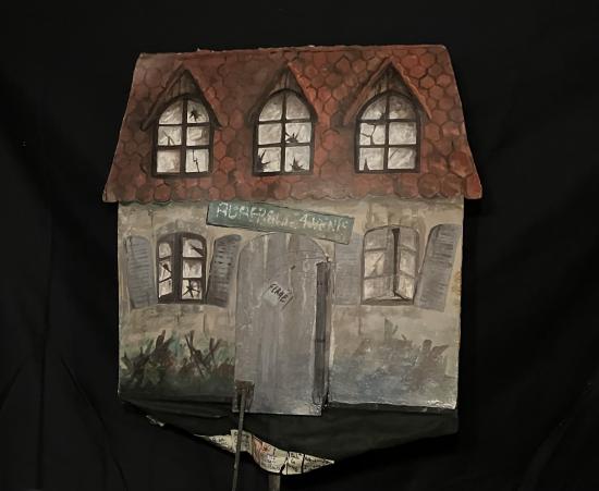 Maison grise à toit brun et fenêtres cassées, un pancarte porte l'inscription "L'Auberge aux 4 vents"