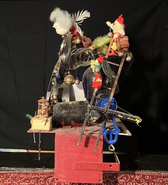 Machine avec rouages, horloge, lutins miniatures, fils, boîtes, le tout sur un socle rouge. Vue du côté droit