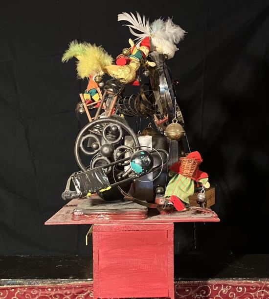 Machine avec rouages, horloge, lutins miniatures, fils, boîtes, le tout sur un socle rouge. Vue du côté gauche