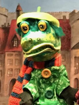 Crapouillard, à tête de crapaud vert avec une écharpe orange et bleue et un béret jaune, devant une place de bourg