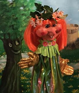 Jeune fille rousse au nez pointu vêtue d'une robe vert pâle et coiffée de fleurs; à côté d'elle, un arbre souriant