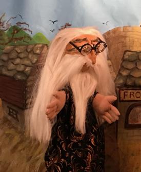 Vieil homme aux cheveux, moustache et barbe blancs, vêtu d'une robe noire à motifs dorés, il porte de grosses lunettes rondes