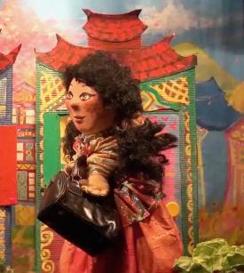 Lilou aux longs cheveux bruns, vêtue d'une robe orange, elle porte une sacoche, on voit une maison à l'arrière-plan