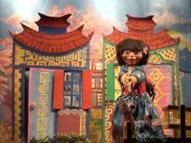 Jeune fille aux longs cheveux bruns, vêtue d'une robe bleue avec une ceinture brune et dorée, maisons à l'arrière-plan