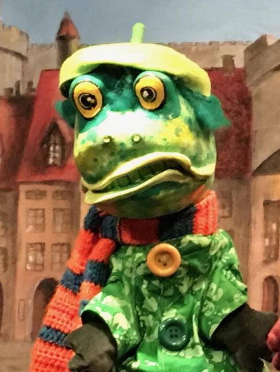 Crapouillard, à tête de crapaud vert avec une écharpe orange et bleue et un béret jaune, devant une place de bourg