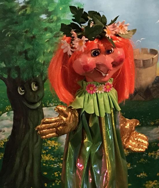 Jeune fille rousse au nez pointu vêtue d'une robe vert pâle et coiffée de fleurs; à côté d'elle, un arbre souriant