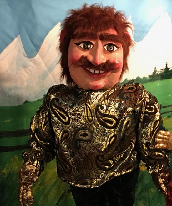 Homme moustachu roux, vêtu d'un pantalon noir et d'une veste dorée à motifs, ses mains dont dorées