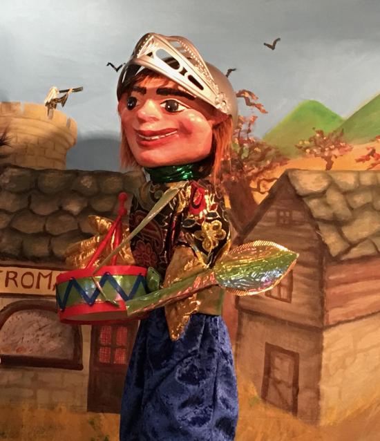 Jeune homme brun aux doigts d'or, vêtu d'un pantalon bleu et d'une veste bariolée, coiffé d'un casque médiéval, il porte un tambour