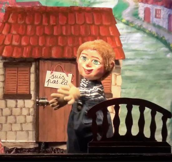 Garçon roux clair aux yeux bleus, vêtu d'une salopette grise et d'un pull à rayures, devant une maison fermée