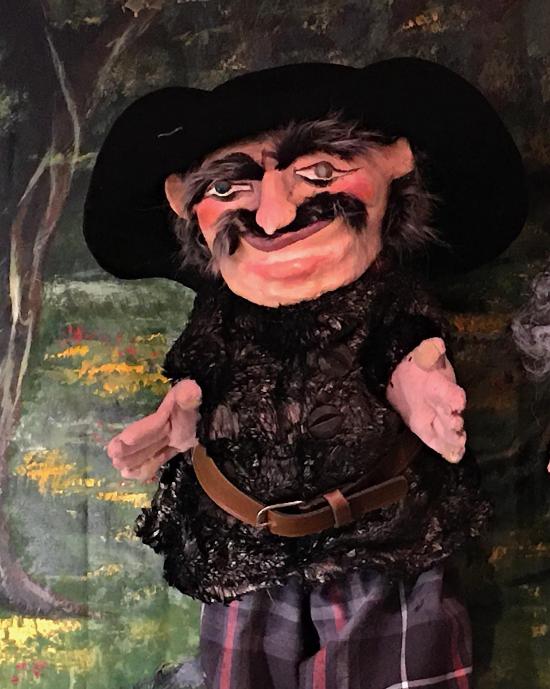 Vieil homme noiraud avec une moustache et des sourcils épais, vêtu d'un pantalon à carreaux et d'une veste noire, coiffé d'un chapeau à larges bords noir