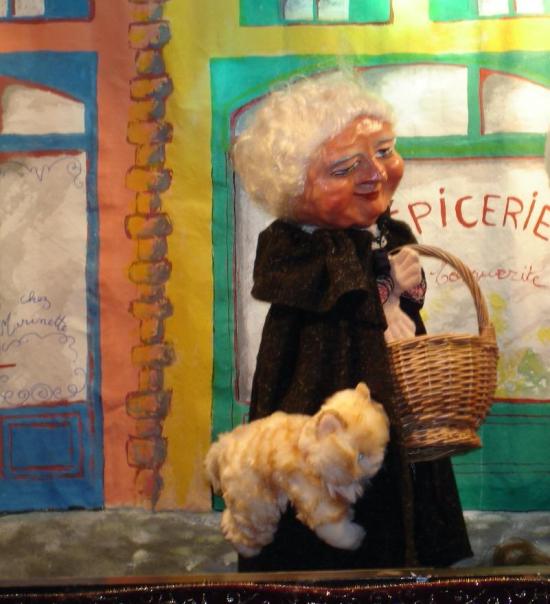 Grand-mère aux cheveux blancs, vêtue d'un long manteau brun, devant une devanture d'épicerie; elle tient un panier au bras et est accompagnée d'un chat