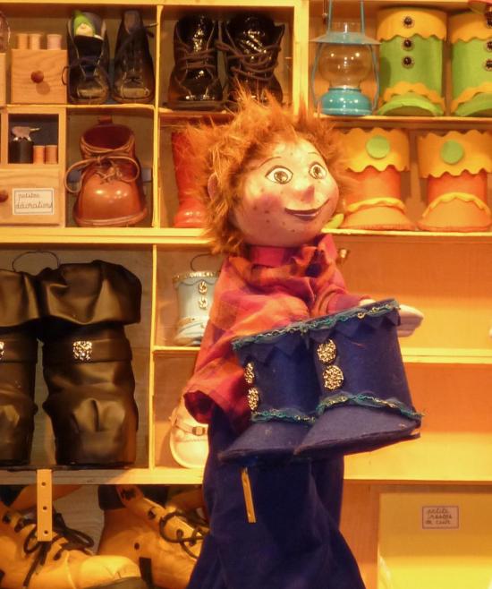 Théo dans la cordonnerie, devant un étal de chaussures, avec une paire de bottes bleues dans les mains