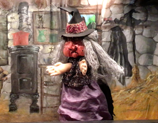 Sorcière au visage rougeaud et longs cheveux blancs, vêtue d'une robe violette, coiffée d'un chapeau pointu gris, dans sa cuisine