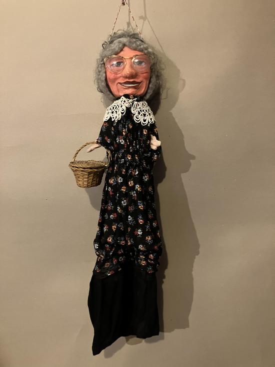 Vieille femme à lunettes, vêtue d'un pantalon noir et d'une tunique à fleurs sur fond noir; elle porte un panier