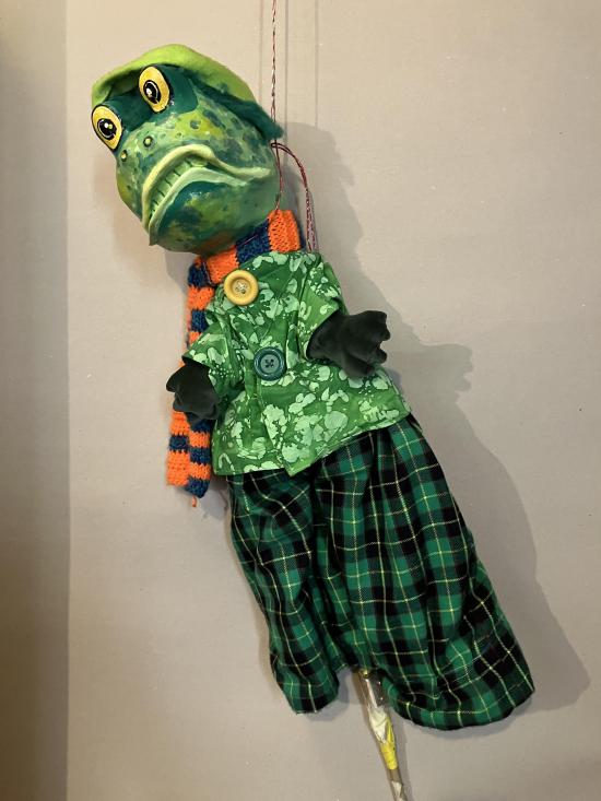 Crapaud vert vêtu d'un pantalon à carreaux vert, d'une veste verte et d'une écharpe orange et bleue