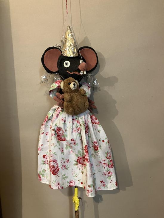 Fée souris vêtue d'une robe à fleurs blanche et rose et d'un tutu doré; elle porte un ours en peluche dans ses bras