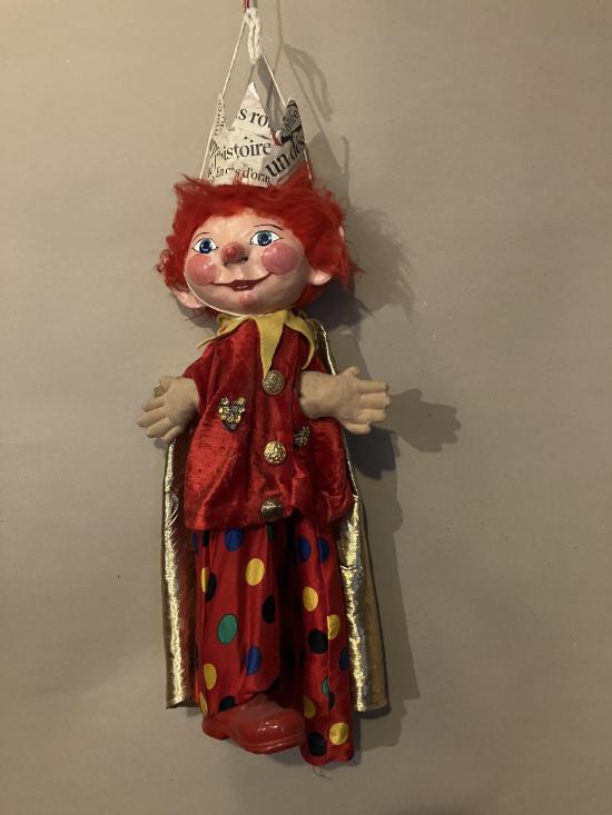 Enfant aux cheveux et pommettes rouges, vêtu d'un pantalon rouge à pois, d'une veste rouge et d'une cape dorée