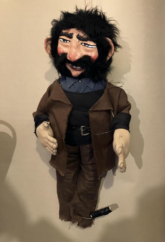 Homme noiraud, barbu et moustachu, vêtu d'un veste et d'un pantalon bruns, avec un couteau à sa cheville