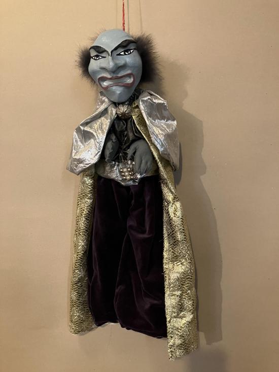 Vieil homme au visage gris, avec une calvitie, vêtu d'une costume brun foncé et d'une longue cape dorée