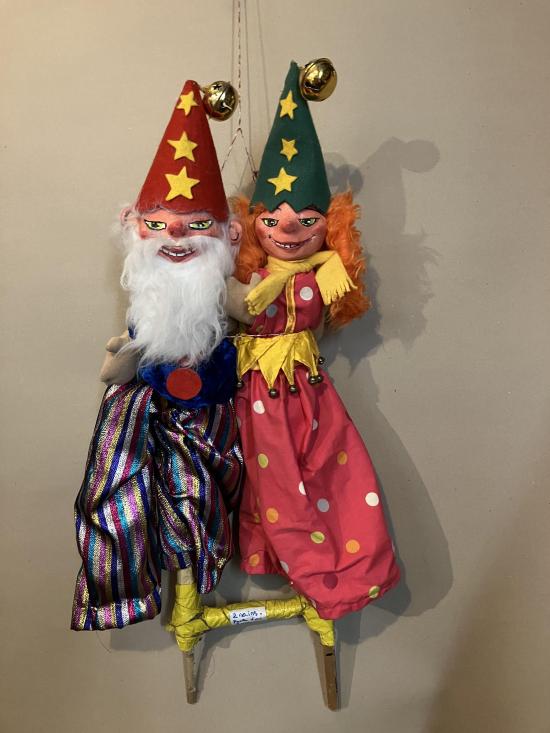 Vieux lutin à barbe blanche et pantalon à rayures de différentes couleurs, coiffé d'un chapeau pointu rouge, jeune lutine rousse à robe rouge et écharpe jaune coiffée d'un chapeau pointu vert