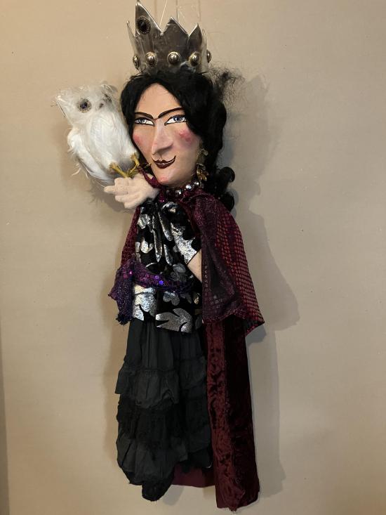 Femme aux longs cheveux noirs, vêtue d'une jupe noire, d'une blouse noire à motifs argentés et d'une longue cape bordeaux, coiffée d'une couronne; elle tient un hibou blanc sur sa main