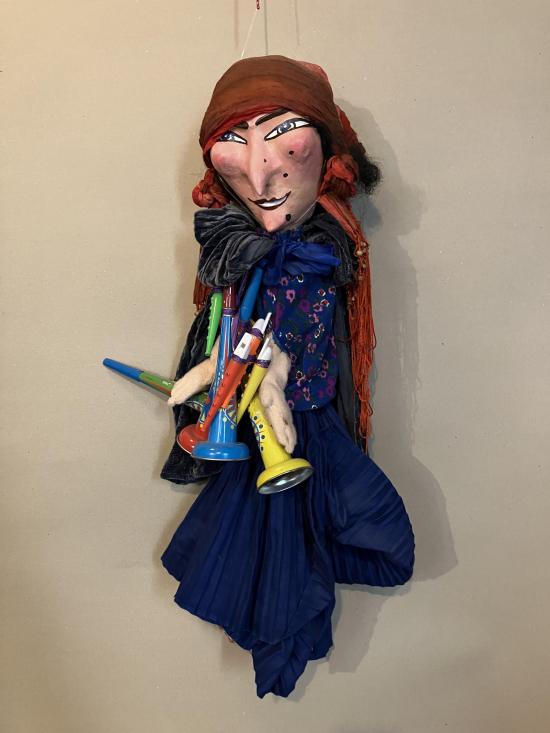 Femme vêtue d'une robe bleue foncé et d'une blouse bleue foncé à fleurs, coiffée d'un turban bordeaux; elle porte de petites trompettes en plastique