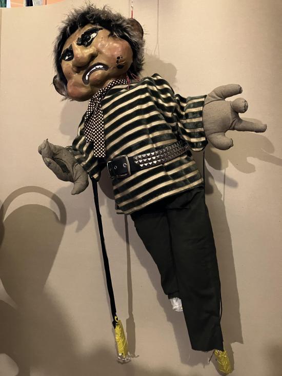 Homme brun aux yeux noirs et à la bouche grimaçante, vêtu d'un pantalon noir, d'un pull rayé noir et blanc et d'une écharpe à petits carreaux noirs et blancs