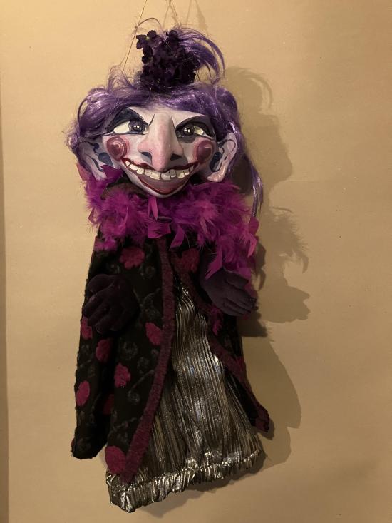 Femme aux cheveux mauves et au sourire menaçant, vêtue d'une robe argentée et d'une cape noire avec des fleurs mauves