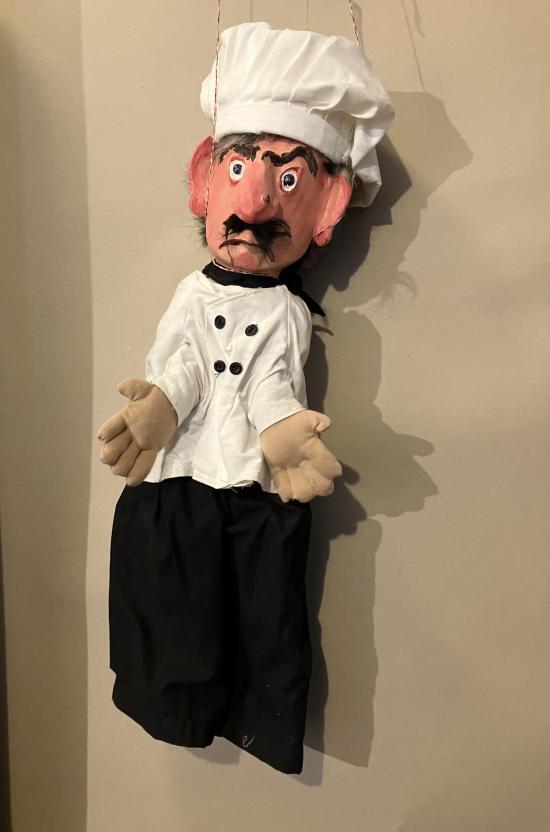 Homme brun moustachu, avec une verrue sur le nez et de gros sourcils, vêtu d'un pantalon voir et d'une chemise blanche de cuisinier, coiffé d'une toque de cuisinier blanche