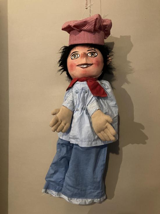 Jeune homme brun aux yeux bruns, vêtu d'un pantalon bleu clair et d'une chemise à petits carreaux bleus et blancs, coiffé d'une toque de cuisinier rose