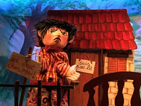 Tibougon devant sa maison, avec une pancarte "Suis pas là" sur sa porte et une autre "Ici c'est chez moi" dans la main