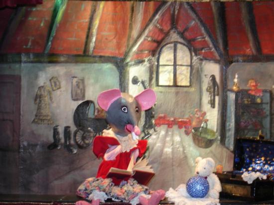 Souris grise au nez et aux oreilles roses, vêtue d'une robe rouge à col en dentelle, dans son grenier avec un livre dans les mains