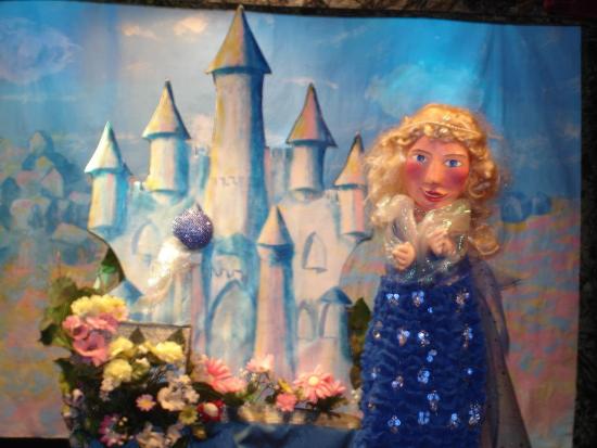 La fée Bellazure, jeune fille blonde vêtue d'une robe bleue, devant son château et une fontaine fleurie