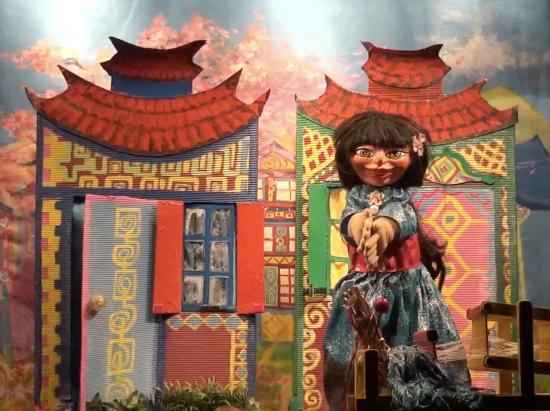 Fleurétoile en Perle d'Or vêtue d'une robe de soie bleue, à côté d'un métier à tisser et devant des maisons colorées