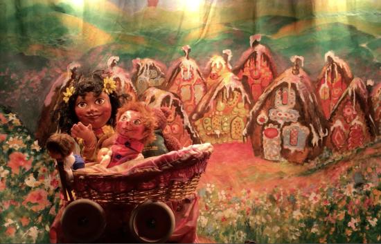 Théo bébé dans le berceau de la fillette, qui se tient debout à côté; en arrière-plan, le village en chocolat, crème et gâteau