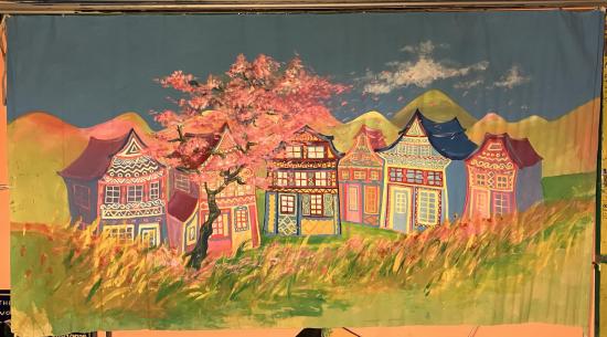 Maisons colorées, collines jaunes à l'horizon, arbre en fleurs rose, pré avec des herbes folles et des fleurs