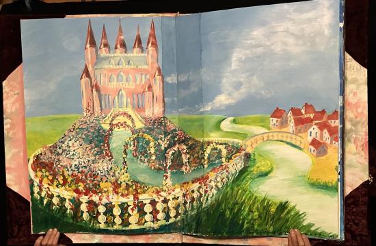 Château rose avec un jardin fleuri devant, sur la droite une rivière et un pont qui le relie à un village